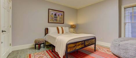 Un dormitorio con cama, mesita de noche, silla, alfombra, cuadro y ventana.