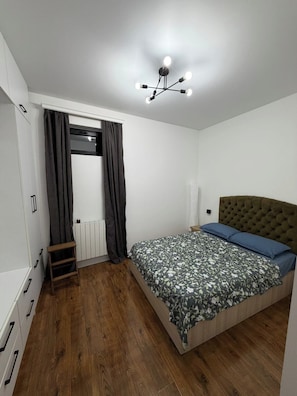 Un dormitorio con cama, radiador, suelo de madera, armario y lámpara en el techo.
