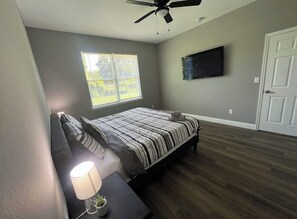 Ein Schlafzimmer mit einem Bett, einem Deckenventilator, einem an der Wand befestigten Fernseher und einem Fenster mit Jalousien.