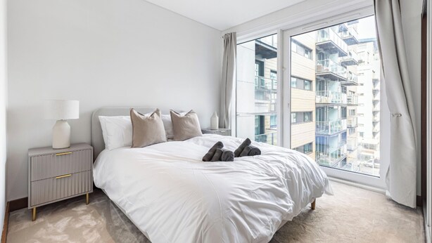 Ein modernes Schlafzimmer mit einem großen Bett, einem Nachttisch und Blick auf eine Stadtlandschaft durch ein großes Fenster.