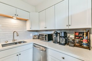 A modern kitchen with white cabinets, a stainless steel dishwasher, a microwave, a coffee maker, and a coffee grinder. - Walk to Beach & Pier Pet-Friendly + Coffee/Tea Bar (Galveston)