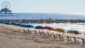 Ein Strand mit Reihen von Liegestühlen und Sonnenschirmen, ein Steg mit einem Riesenrad und das Meer im Hintergrund.