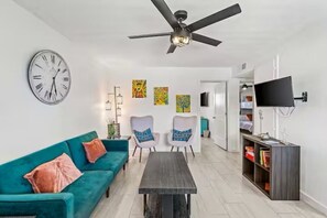 Ein Wohnzimmer mit einem türkisfarbenen Sofa, einem Couchtisch, zwei Sesseln, einem Deckenventilator, einer Wanduhr und einem Fernseher.