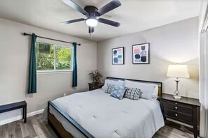 Ein Schlafzimmer mit Bett, Deckenventilator, Fenster mit Vorhängen, Nachttisch mit Lampe und zwei gerahmten Bildern an der Wand.