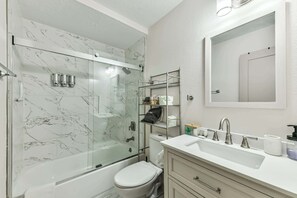 Casa de banho com chuveiro em cabine de vidro, sanita branca, lavatório com espelho e prateleira com produtos de higiene pessoal.