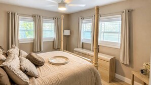 Une chambre à coucher avec un lit à baldaquin, deux fenêtres avec des rideaux, une table de chevet avec une lampe et une petite table sur laquelle est posé un vase.