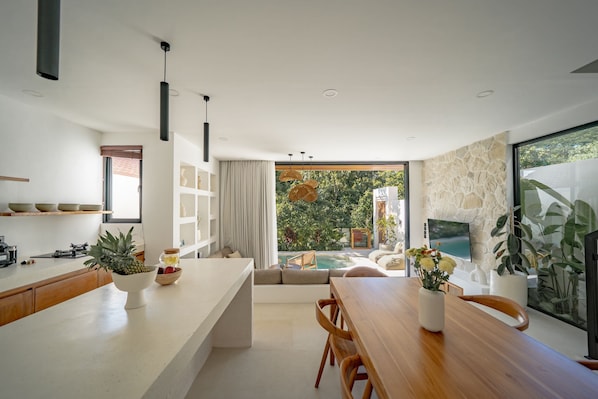 A modern kitchen with a large island, a dining area with a wooden table, and a view of a swimming pool and greenery. - Villa Alam Indah Uluwatu (Uluwatu)