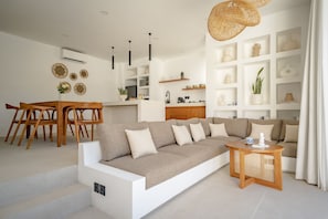 Una sala de estar moderna con un sofá, una mesa de comedor de madera y sillas. Se observan estanterías con objetos decorativos y una cocina al fondo.