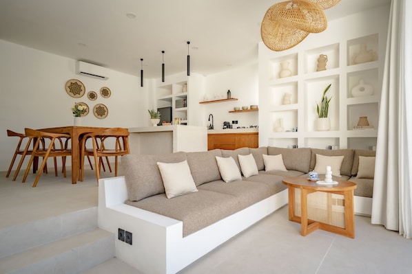 Ein modernes Wohnzimmer mit einer Couch, einem Holzessttisch und Stühlen. Im Hintergrund sind Regale mit Dekoration und eine Küchenzeile zu sehen.