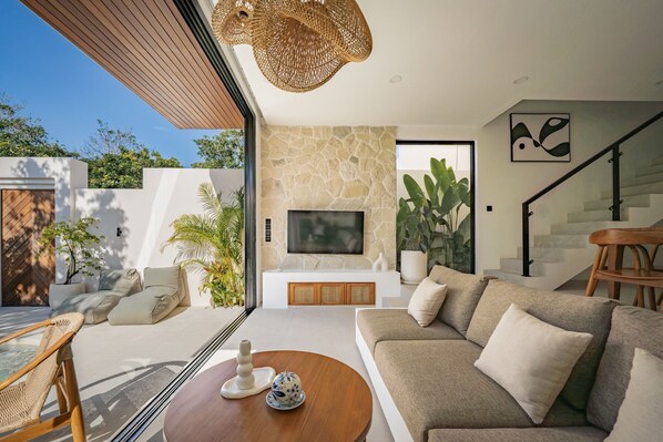 A modern living room with a stone wall, a flat-screen TV, a sofa, a coffee table, and a wicker chair. - Villa Alam Indah Uluwatu (Uluwatu)