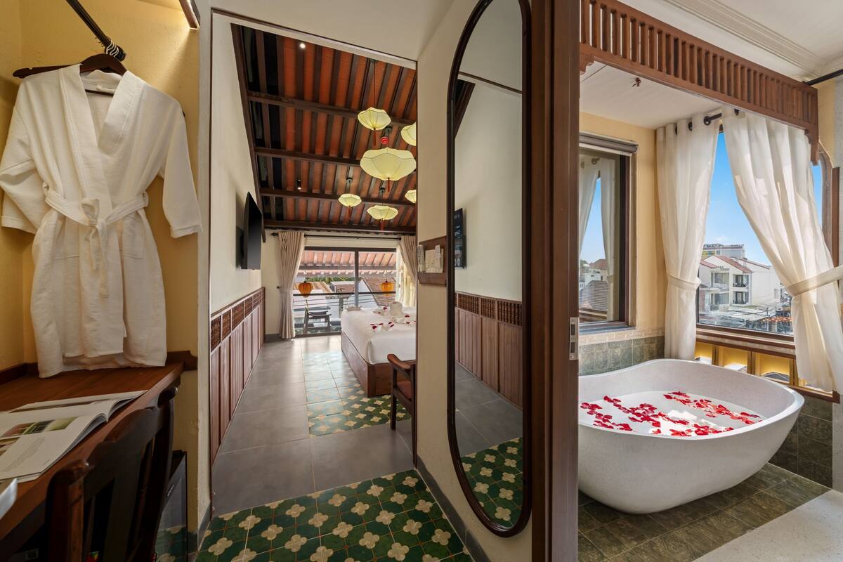 A hotel room with a large bathtub, a wooden ceiling, a view through a window, and a hallway leading to a dining area.