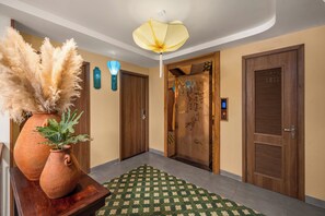 Un couloir avec un tapis à motifs verts, un vase contenant des plantes séchées, une plaque numérotée fixée au mur et une porte en bois dotée d’un panneau à persiennes.