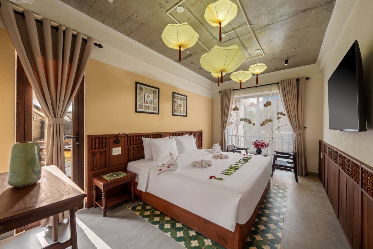 A hotel room with a bed, wooden headboard, bedside tables, a green vase, a wooden bench, a TV, and a window with curtains.