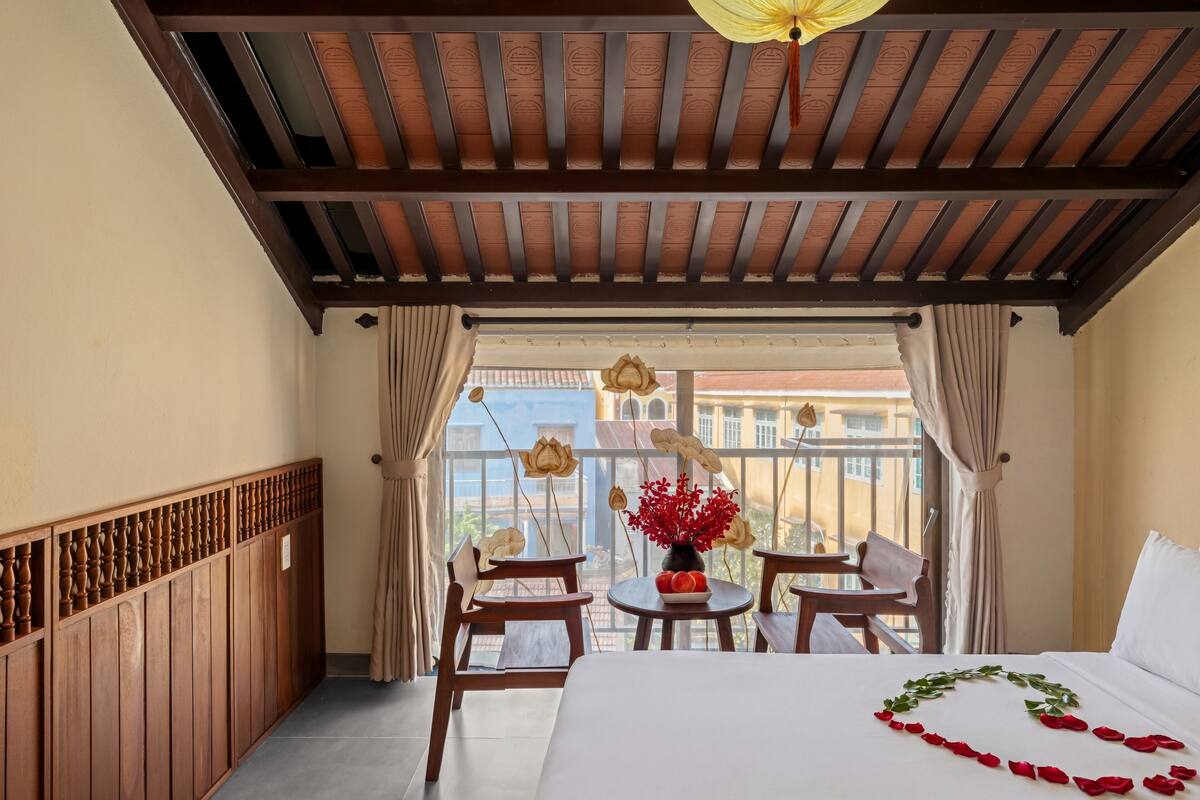 A hotel room with a balcony, a bed with rose petals, a table with a vase, and a view of buildings.