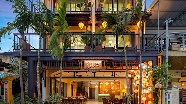 A hotel with a traditional design, featuring a balcony with plants, a reception area with a sign, and a dining area with tables and chairs.