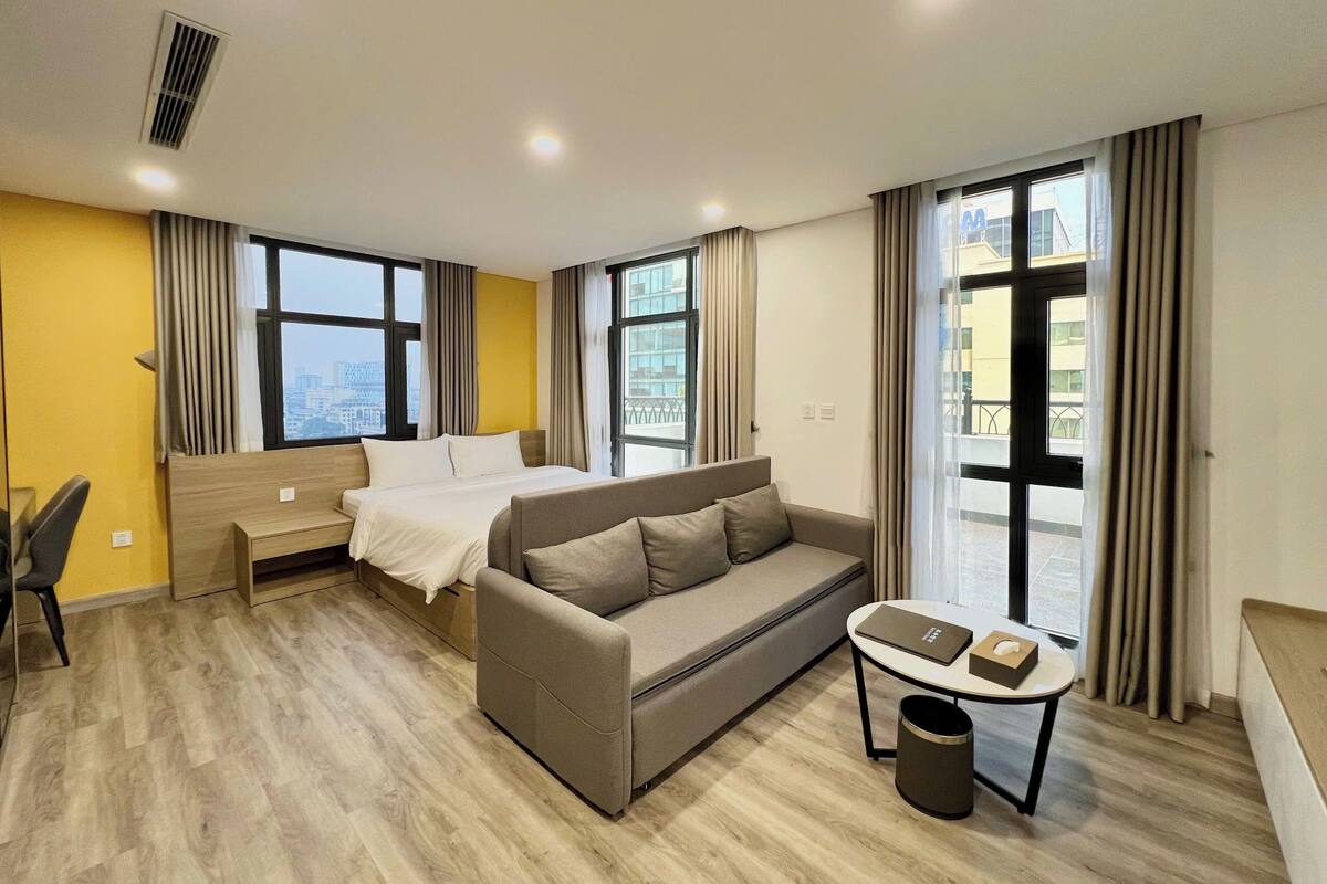 A hotel room with a bed, sofa, desk, and large windows.