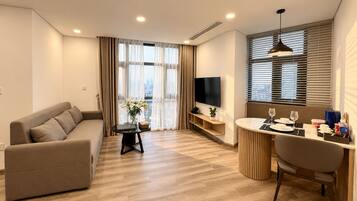 A modern living room with a sofa, a dining table with chairs, a TV, and a large window with blinds.