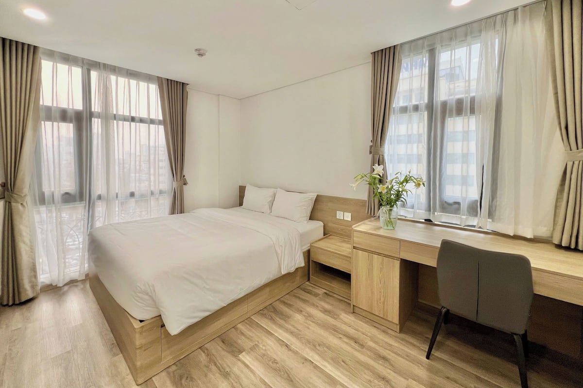 A hotel room with a bed, desk, chair, and large windows with curtains.