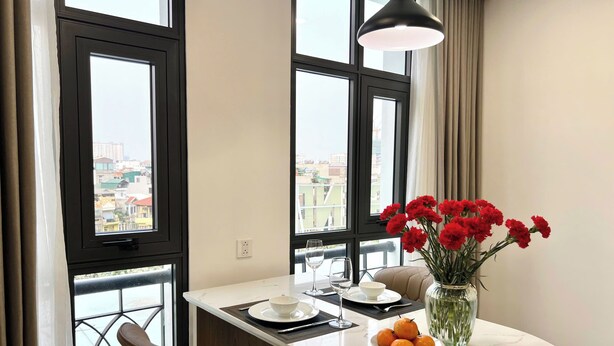 Un espace de repas moderne comprenant une table, des chaises, un vase de fleurs et une vue sur la ville grâce à de grandes fenêtres.