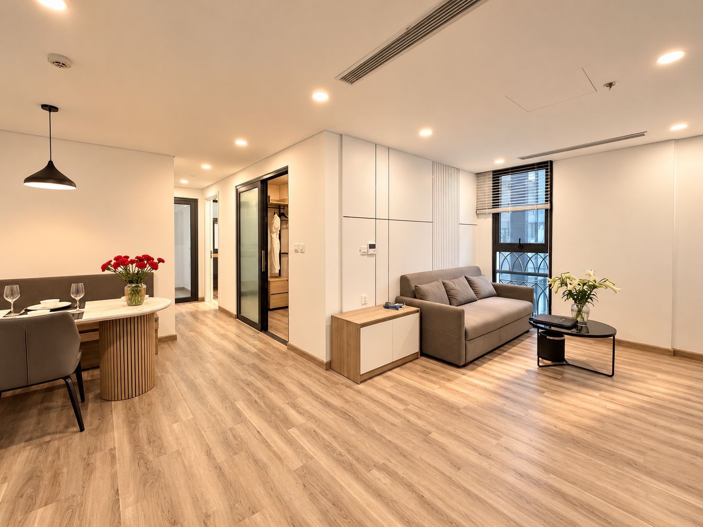 A modern living room with a sofa, a dining table with chairs, a small side table, and a window with blinds.