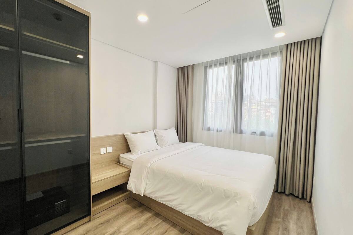 A modern hotel room with a bed, a small bedside table, a built-in wardrobe, and a large window with curtains.