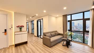 A modern hotel room with a sofa, a small table, a vase with flowers, and a city view through large windows.