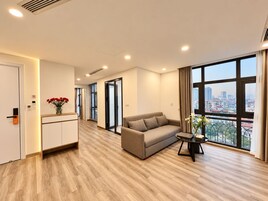 A modern hotel room with a sofa, a small table, a vase with flowers, and a city view through large windows.