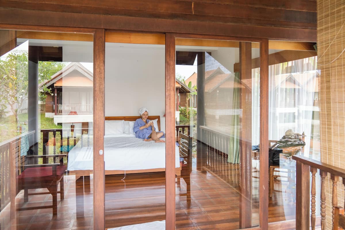 Un balcón de madera con una barandilla de vidrio, una cama con ropa blanca y una vista de una casa al fondo.