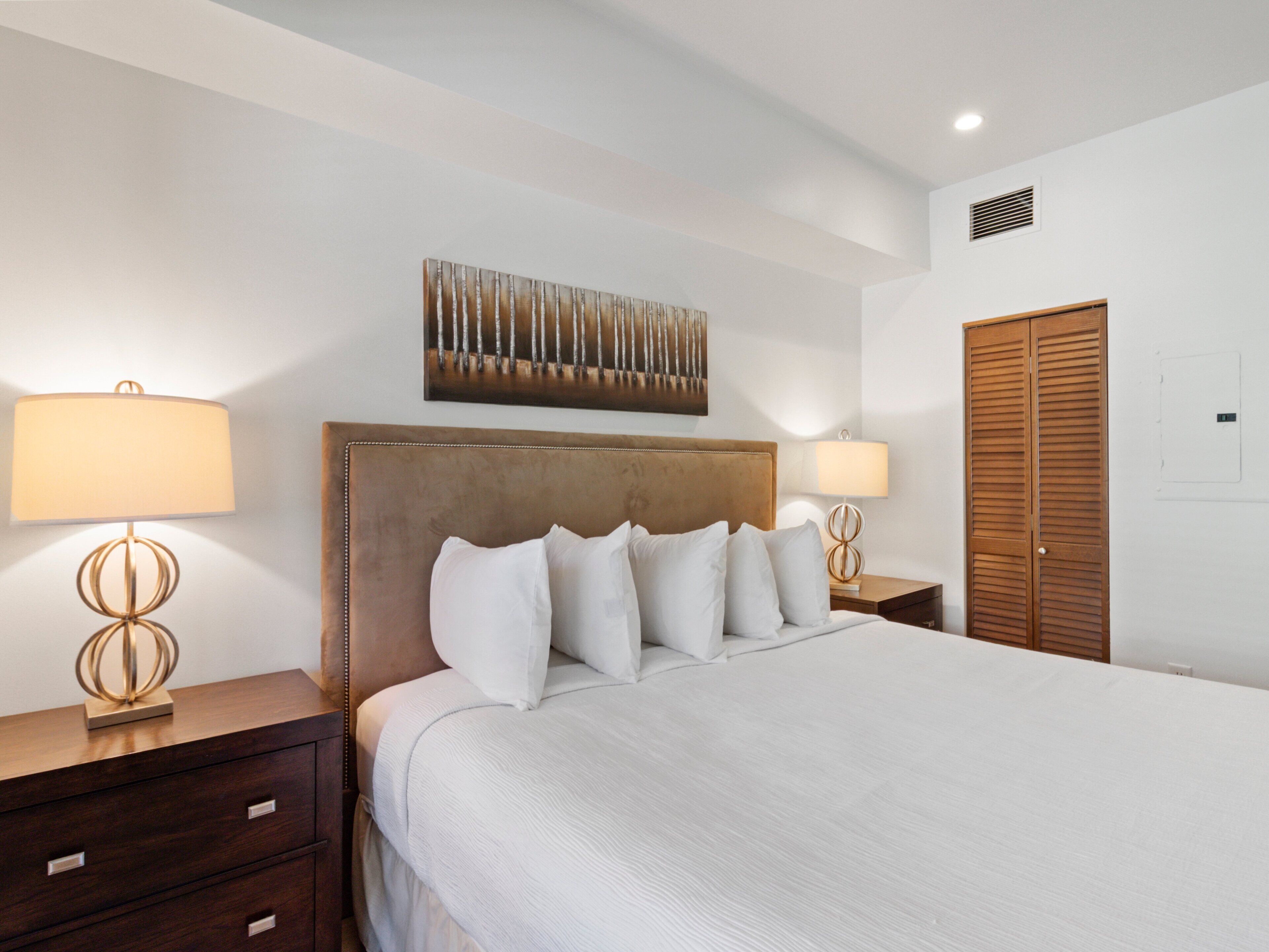 Habitación de hotel ordenada con una cama grande, dos mesitas de noche, un armario de madera y una obra de arte colgada en la pared.