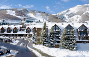 A snow-covered lodge with multiple buildings, a ski lift in the background, and a road with parked cars. - Lodge at Mtn Village A226 | Ski-In/Out | Hot Tub | Plaza View | PCMR | Pool (Park City)
