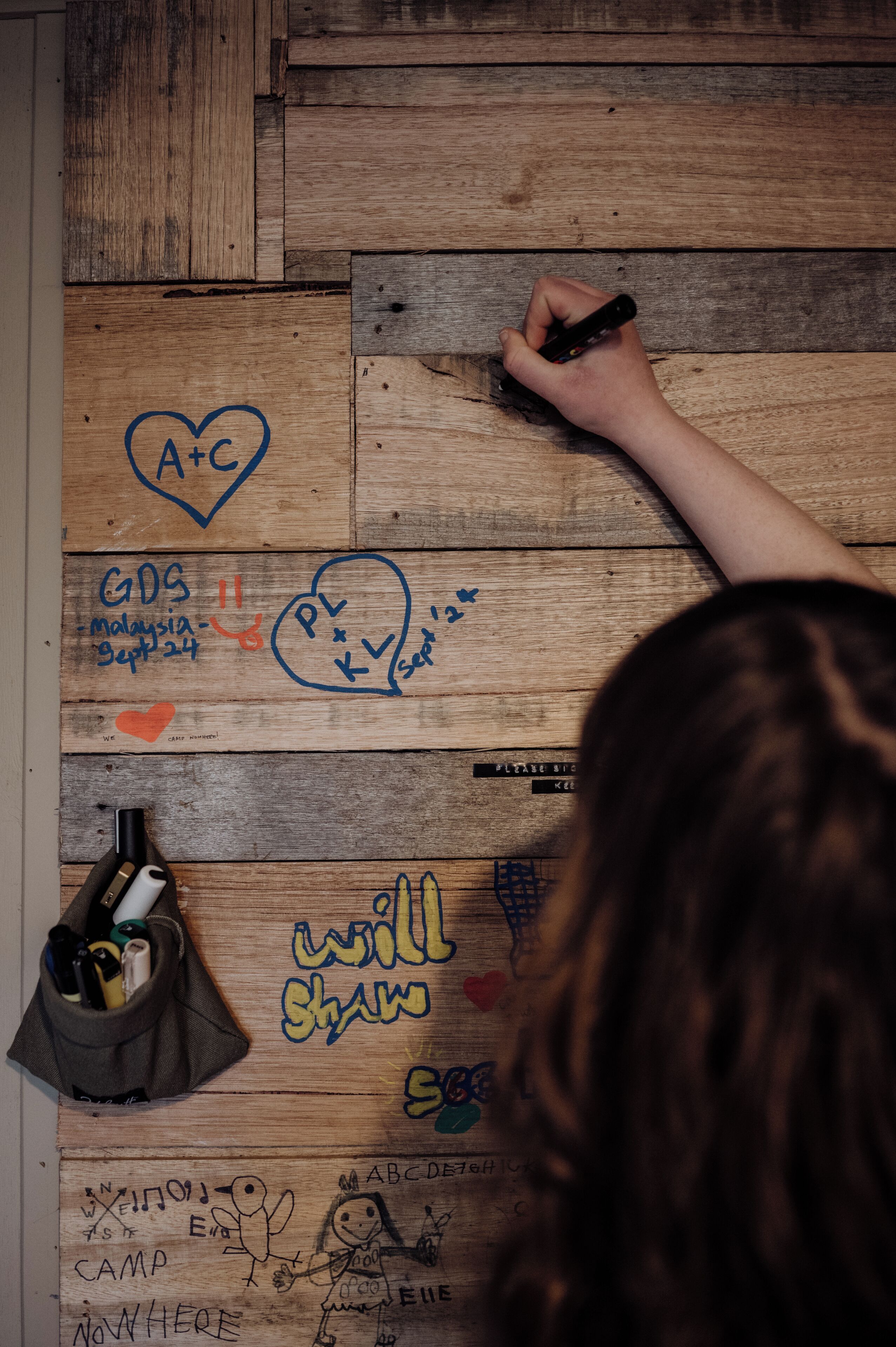 Una mano scrive su una superficie di legno con diversi disegni e testi, tra cui 'A+C', 'GDS Malaysia Sept 24' e 'will shawn'.
