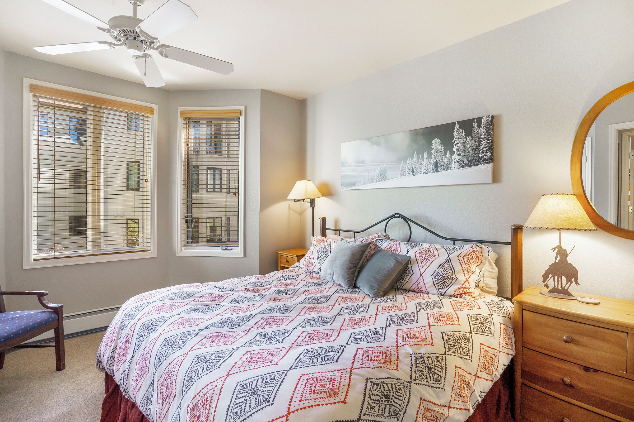 A bedroom with a bed, a nightstand, a lamp, a chair, a ceiling fan, and two windows with blinds.