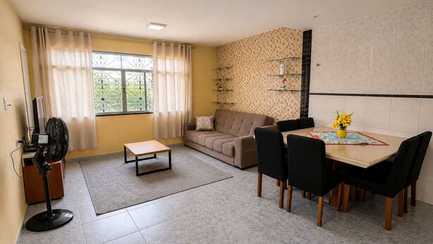 A living room with a sofa, a dining table with chairs, a fan, and a window with curtains. - Casa confortável (Belém)