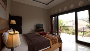 Un dormitorio con cama, televisión, lámpara y balcón con vista a palmeras.