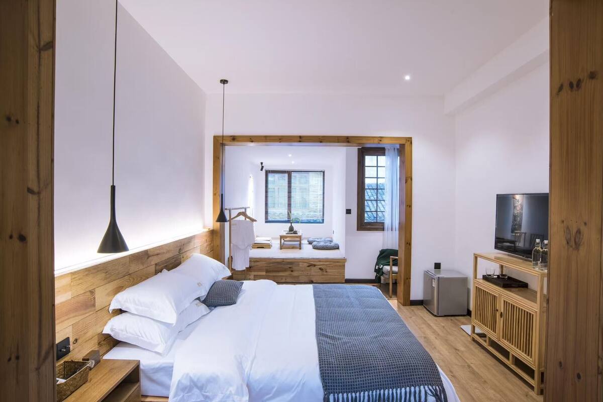 Een slaapkamer met een bed, nachtkastje, televisie en uitzicht op een kamer met een raam.