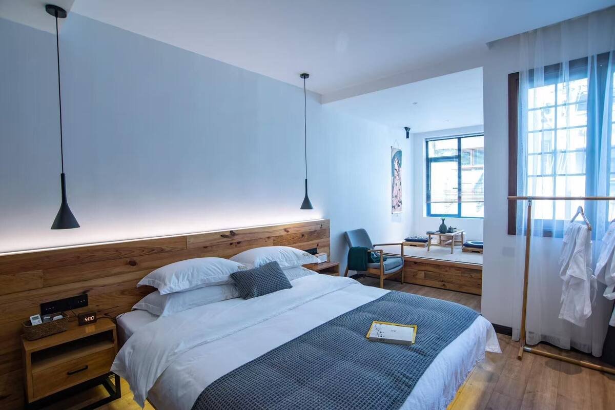 Een moderne hotelkamer met een houten hoofdbord, een bed met wit en grijs beddengoed, een nachtkastje met een lamp, een stoel, en een bureau met een stoel.
