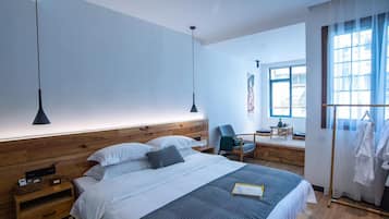 A modern hotel room with a wooden headboard, a bed with white and gray bedding, a bedside table with a lamp, a chair, and a desk with a chair.