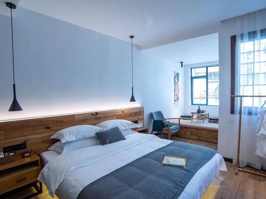 A modern hotel room with a wooden headboard, a bed with white and gray bedding, a bedside table with a lamp, a chair, and a desk with a chair.