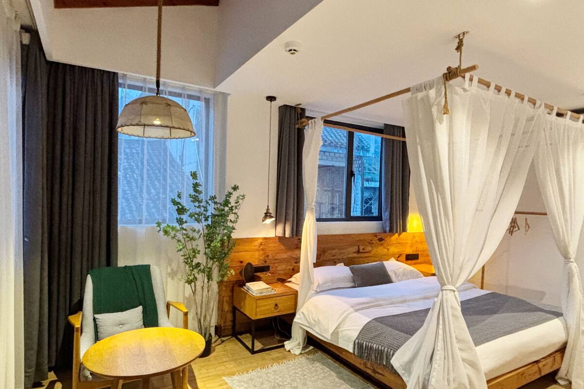 Een slaapkamer met een bed, nachtkastje, lamp, stoel en uitzicht op een gebouw.