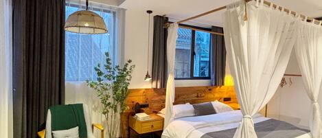 Un dormitorio con cama, mesita de noche, lámpara, silla y vista a un edificio desde la ventana.