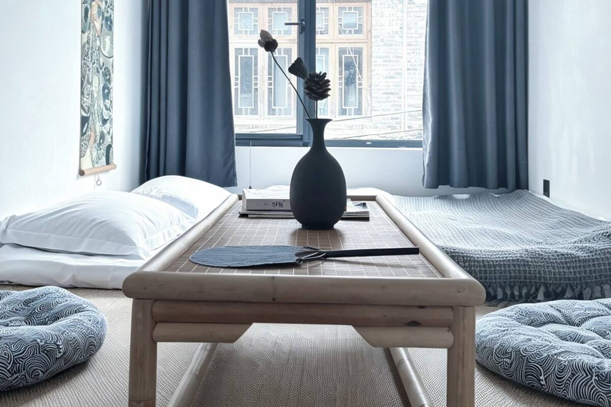 Een kamer met een bed, een klein tafeltje, een raam met gordijnen, een schilderij aan de muur en uitzicht op gebouwen.