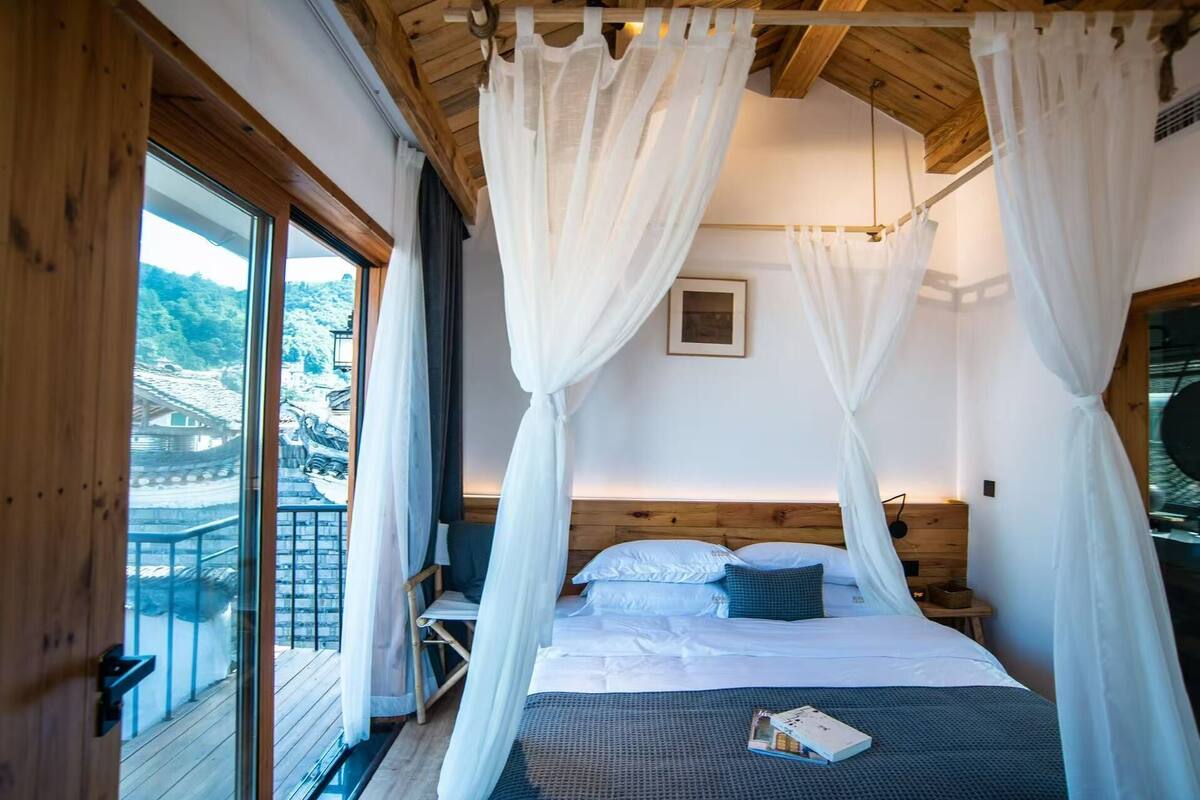 Een slaapkamer met een bed, witte gordijnen, een houten hoofdbord, een klein nachtkastje en uitzicht op een traditioneel gebouw.