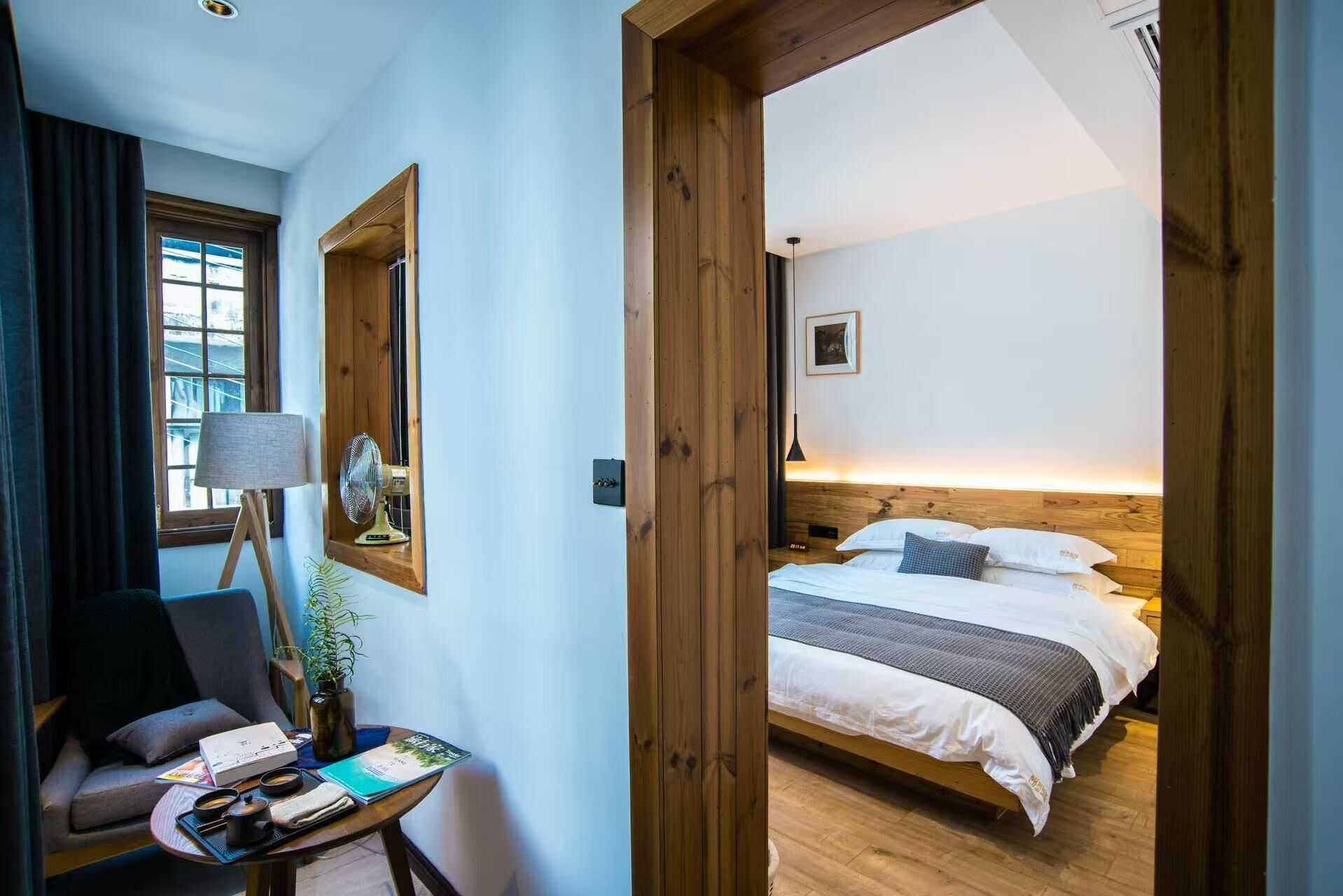 Habitació d'hotel amb un llit, una cadira, una taula petita amb una planta i una finestra amb cortines.