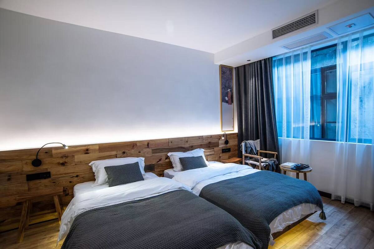 Een hotelkamer met twee bedden, een houten hoofdeinde, een raam met gordijnen, een stoel en een wandlamp.