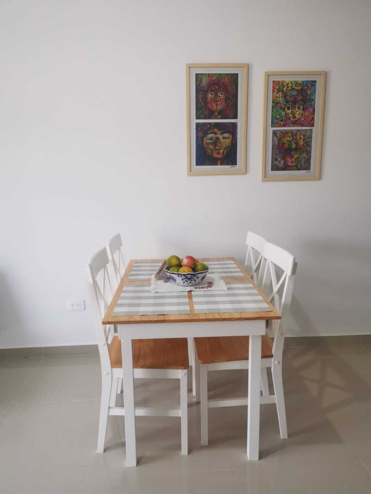 Une table à manger avec un saladier de fruits, des chaises blanches et deux œuvres d’art encadrées accrochées au mur.