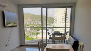 Een moderne woonkamer met een eethoek, grote ramen, een flatscreen televisie en uitzicht op een stadsgezicht.