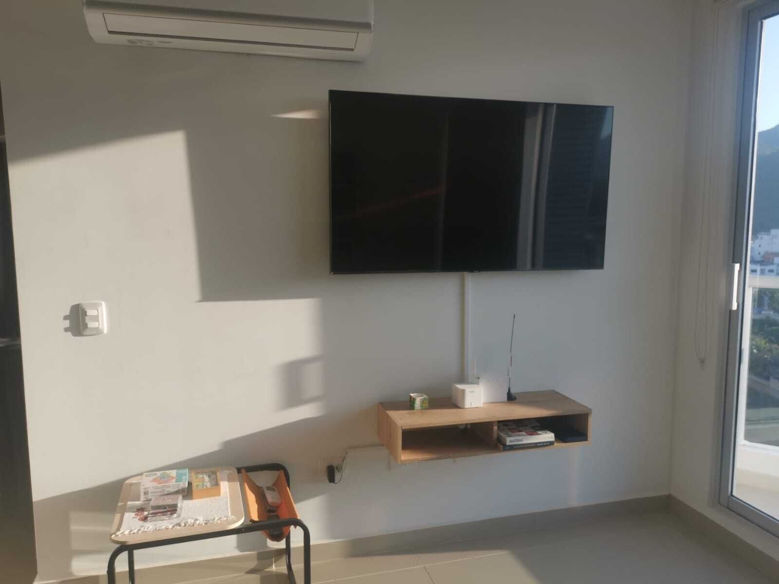 A room with a mounted TV, a small wooden shelf, a desk with a chair, and a window.