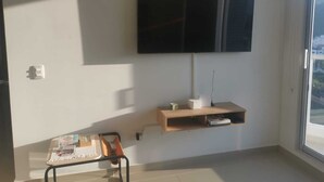 Een kamer met een wandtelevisie, een klein houten plankje, een bureau met een stoel, en een raam.
