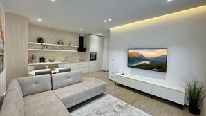 Een moderne woonkamer met een hoekbank, een flatscreen televisie aan de muur en een keuken op de achtergrond.