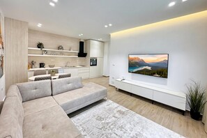 Ein modernes Wohnzimmer mit einer Eckcouch, einem Flachbildfernseher an der Wand und einer Küchenzeile mit weißen Schränken und einer schwarzen Dunstabzugshaube.